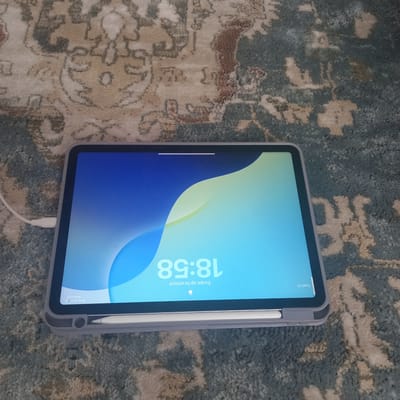 The image shows a tablet device with a protective grey case, lying on a carpet with a floral pattern in muted tones of beige, brown, and teal. The tablet's screen displays a wallpaper with a gradient from deep blue to light blue and a soft yellow-green wave. The time displayed on the lock screen is 18:58, with the text "Swipe up to unlock" below it. A charging cable is plugged into the left side of the tablet, with the cable extending off-frame to the left. A stylus is magnetically attached to the bottom edge of the tablet. The scene is static and there are no people present. The environment appears to be indoors, likely a home setting given the carpet. There is no indication of the location within the city of Cepeleuți, Moldova.