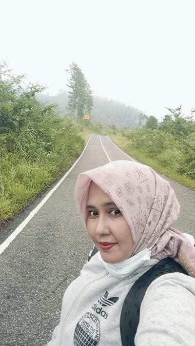 A low-angle, eye-level medium shot captures a woman taking a selfie on a paved road that recedes into a foggy landscape. The woman, wearing a pink hijab and a gray hooded sweatshirt with the Adidas logo and "World Brand with the 3 Stripes" text, is positioned on the right side of the frame, with her face close to the camera. She has dark hair peeking from under her hijab and is wearing red lipstick. A white face mask is pulled down below her chin. The road, with a white dividing line, is flanked by lush green grass and vegetation on the left and a gentler slope of greenery on the right, leading up to a misty forest. Tall, thin trees stand on the right side of the road, partially obscured by fog. A yellow rectangular sign is visible further up the road on the right. The overall atmosphere is overcast and hazy, suggesting a cool, damp environment in Bintangpermata, Indonesia.