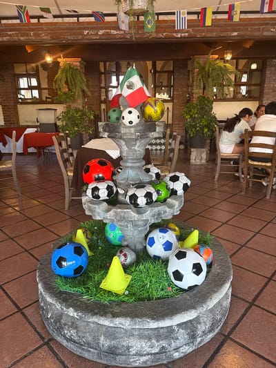 A medium shot captures an indoor dining area with a complex array of decorative elements. A prominent centerpiece is a weathered gray stone planter overflowing with artificial green grass and an assortment of soccer balls in various colors and designs. More soccer balls are arranged in a stone fountain alongside small, yellow plastic cones.

International flags of various nations hang vertically along the walls, adding to the global theme. In the background, several people are seated at tables, one woman with long dark hair is facing away from the camera. The restaurant's architectural style features brick walls and dark wood framing, creating a warm ambiance. The floor is tiled, and the lighting is soft and diffused.

The scene is located in Tepeji de Ocampo, Mexico.