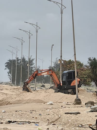 The image displays a sandy area, likely a beach or shoreline, in Thuận An Ward, Vietnam, under an overcast sky. A compact orange and black excavator is situated in the midground, partially obscured by accumulated sand, with its bucket embedded in a large sand mound. Behind the excavator, a row of tall, light-colored streetlights with curved, decorative arms extends into the background, flanked by a line of dark green trees with dense foliage. A body of water, likely the ocean, is visible on the far left horizon. The foreground consists of light-colored sand extensively scattered with various debris, including plastic items, organic matter, and fragments of rock. No individuals are present.