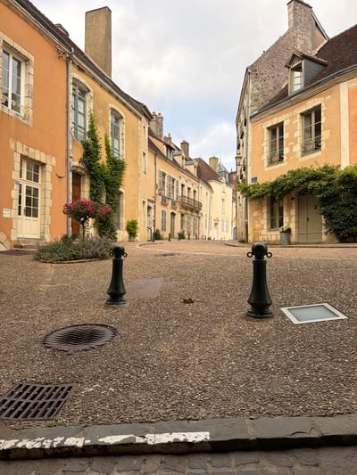 The image depicts a cobblestone street in Bellême, France, with traditional European architecture lining both sides. The buildings are a mix of light orange and yellow hues, with stone accents around windows and doors. Green ivy climbs up some of the facades, and potted plants add a touch of color. In the foreground, two black bollards stand on the street, and a manhole cover and a drain are visible on the ground. The sky is overcast, suggesting a cloudy day. The street appears quiet and empty, with no people or vehicles present.