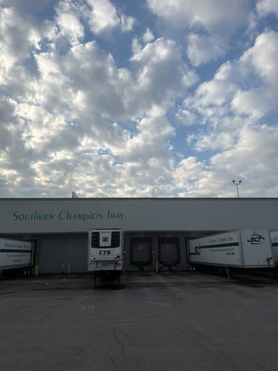The image captures a loading dock area of a commercial building with "SOUTHERN CHAMPION TRAY" prominently displayed on its facade. Several semi-trailers are parked at the loading bays, identified by numbers 34, 36, and 37. One trailer displays the text "SOUTHERN CHAMPION TRAY, LP" and a website address "sctray.com". Another trailer has a "CTS" logo and "THERMO KING" branding, indicating it's a refrigerated unit. The environment appears to be an industrial or logistics facility. The sky above is partly cloudy with dramatic cloud formations. The ground is asphalt, with painted numbers "35", "36", and "37" visible in yellow. The scene suggests a business operation involving shipping and receiving. This facility is located in Chattanooga, United States of America.