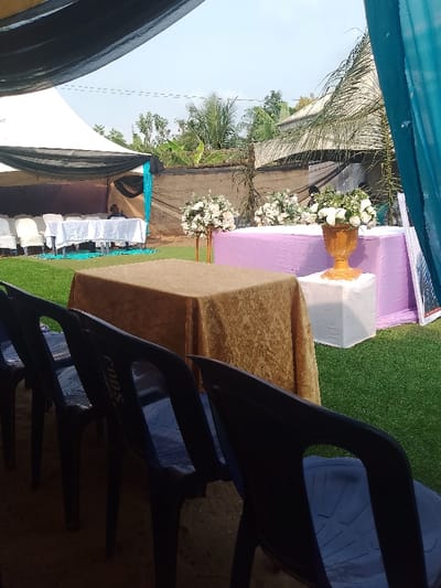 The image displays an outdoor event setup in Ufuma, Nigeria, under clear daylight conditions. The ground is covered with green artificial turf. In the foreground, four dark plastic chairs are arranged in a row, oriented away from the camera. Behind these, a rectangular table draped with a gold-patterned tablecloth is positioned. To its right, a larger rectangular table is covered with a solid lavender tablecloth. This lavender table holds two white floral arrangements in gold urns; one urn is elevated on a white cube-shaped pedestal. A third white floral arrangement on a taller, slender gold stand is situated to the left of the lavender table.

In the mid-ground, a white canopy tent is visible, partially draped with black fabric and accented with a section of teal fabric. Beneath this tent, multiple white tables with white tablecloths are arranged, surrounded by white chairs. One individual, wearing a dark top, is seated at a white table under the tent, facing away from the viewer. The background features a concrete or mud wall, dense green vegetation including palm trees and banana plants, and glimpses of distant structures. A teal fabric drape hangs vertically on the right side of the frame, and dark fabric covers the upper portion of the immediate foreground, suggesting an overhead covering providing shade.