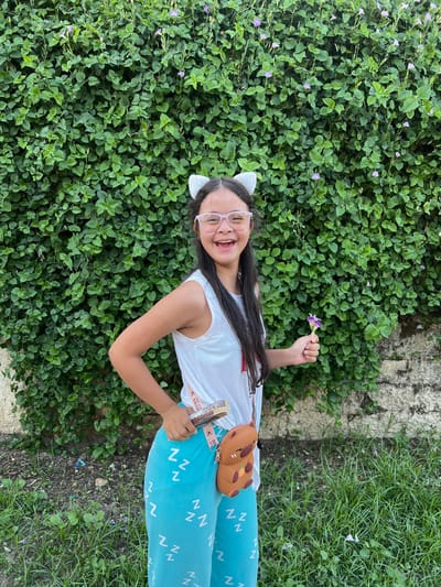A young person with Down syndrome stands outdoors in front of a lush green wall of foliage. The individual is wearing cat ears, glasses, a white tank top, and teal pants patterned with white "Z"s. They are holding a small purple flower in their right hand and a candy bar in their left. A brown bear-shaped purse is slung across their body. The background consists of a dense, leafy plant with small purple flowers and a partial view of a stone wall and grassy ground. The scene appears to be captured during daylight, with the sun casting a soft light. This image was taken in Villa de Cura, Venezuela.
