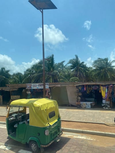 The image depicts a street scene in Djondji, Benin. A green and yellow auto rickshaw is parked on the right side of the image, adjacent to a paved walkway. Beside the walkway is a sandy area leading to a row of shops or stalls, constructed with rustic materials like wood and corrugated metal. These stalls display various goods for sale, including textiles, handicrafts, and tourist souvenirs. Palm trees are visible behind the stalls, reaching towards a blue sky with scattered clouds. A street sign is on the left side. No people are directly visible in the image, although a figure may be inside the rickshaw. The scene appears to be taken during daylight.