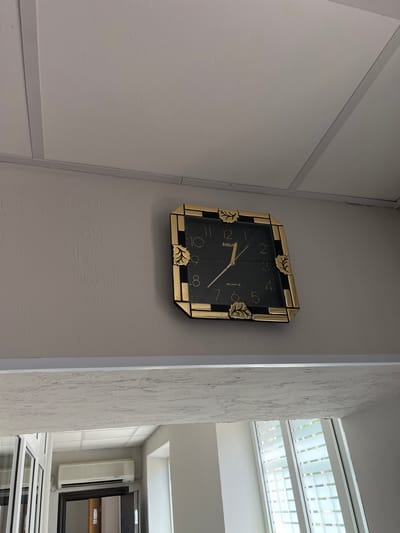 This is a medium shot, slightly low-angle, exterior, daylight, medium-resolution, color photograph of a square clock mounted on a textured, light-colored wall. The clock has a gold border with decorative leaf accents and a black face with gold numbers and hands. The time displayed on the clock is approximately 10:08. The clock is positioned within a room, indicated by the ceiling panels above and a textured beam below. The room appears to be indoors, with a glimpse of an air conditioning unit and a doorway in the background through an open archway. The overall lighting suggests it is daytime, but it is not possible to determine the exact time of day from the image. There are no people or animals visible in the image. The location is identified as Nisporeni, Moldova.