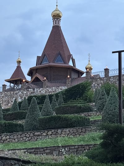 The image shows a close-up, rotated view of a building with onion domes, typical of Russian Orthodox architecture. The building appears to be constructed of wood and stone, with a dark brown roof. Several golden onion domes topped with crosses are visible. The structure is surrounded by lush greenery, including manicured hedges and trees, as well as stone walls. The overall impression is that of a church or monastery, possibly set in a garden or park. The lighting suggests it is either early morning or late evening, with soft, diffused light and a cloudy sky. The image does not contain any people or visible text. The provided location context is Staraya Stanitsa, Russia, which aligns with the architectural style.