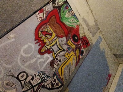 A concrete surface displays vibrant graffiti art. Dominant colors are red, yellow, black, and brown, outlining abstract shapes and figures. A profile with a red arrow through the eye is visible, along with geometric patterns and stylized lettering. The artwork appears spontaneous, utilizing bold lines and contrasting colors to create a striking visual. Some faded white spray paint is near the art. A portion of the artwork extends onto a neighboring, blue-gray surface.