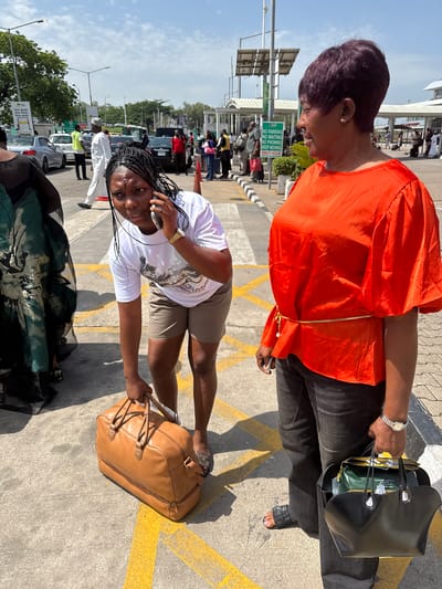 In Gwaska, Nigeria, a young woman with braided hair is on the phone, bending over to pick up a brown leather duffel bag. She is wearing a white t-shirt and khaki shorts. To her right stands another woman with short, dark hair, wearing a bright orange top and black pants. She is holding a black handbag filled with various items, including what appears to be a green box or package. They are standing on a paved area with yellow markings, likely an airport tarmac or parking lot, under a bright, sunny sky with some clouds. In the background, there are several vehicles, including cars and what might be a bus, as well as other people and airport infrastructure. A sign with "NO PARKING NO WAITING NO PHONING KEEP MOVING" is visible in the middle ground.