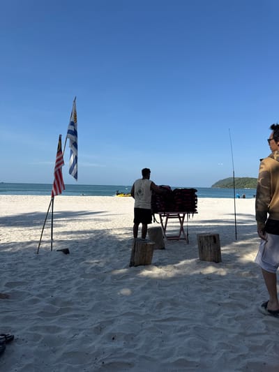 A beach scene under a bright blue sky. White sand covers much of the foreground and stretches to the ocean. Two flags, one appearing to be Malaysian, are planted in the sand. A man stands near a table laden with red life vests. Wooden stumps surround the table. Two people are at the top of the picture, presumably walking by, and a fishing rod stands tall in the sand. The beach appears sunny and inviting.