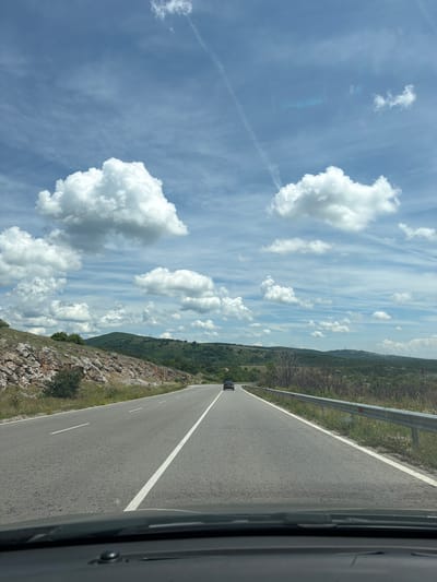 The image, taken from inside a vehicle looking forward, captures a scenic drive on a clear day in Bogyovtsi, Bulgaria. The perspective is from the passenger side, with part of the car's interior visible on the far right.

The central subject is a two-lane asphalt road, stretching into the distance with a solid white line marking the right edge and a broken white line down the center. A dark-colored car is visible ahead, further down the road. To the right of the road, a metal guardrail runs parallel to the asphalt, separating it from the shoulder.

On both sides of the road, the terrain is rugged and hilly, covered with sparse vegetation, rocks, and some patches of green grass. In the background, rolling mountains or large hills are visible, densely covered with trees and greenery, suggesting a rural or mountainous region.

The sky dominates the upper portion of the image, displaying a bright blue hue with numerous white, fluffy cumulus clouds scattered across it. There are also some fainter, wispy cloud formations. A faint contrail from an aircraft is visible in the upper left of the sky. The sunlight indicates it is daytime, likely mid-morning or afternoon, with fair weather conditions. No people are visible, and there is no discernible text in the image.