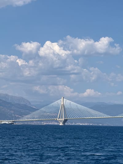 This is a photograph of the Rio–Antirrio Bridge in Greece. The bridge, a prominent suspension bridge, stretches across a body of water that appears to be the Gulf of Corinth. The bridge's elegant design, with its numerous cables fanning out from a central tower, is a central subject. The water is a deep blue, with a textured surface suggesting gentle waves or ripples.
The background features a coastline with mountains. The mountains are muted in color, indicating distance and possibly a hazy atmosphere. The sky above is a bright blue, filled with fluffy white clouds, suggesting fair weather. The lighting indicates it is daytime, likely midday or early afternoon, given the brightness and the shadows.
The scene is serene and vast, with the bridge acting as a man-made structure connecting the natural elements of sea, sky, and land. There are no people visible in the frame. The overall impression is one of architectural achievement within a beautiful natural landscape. The photograph is oriented vertically, with the bridge running from the bottom left to the top right, and the sea occupying the right half of the frame.