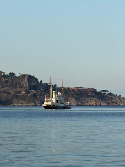 The image shows a sideways view of a body of water, likely the Ionian Sea near Chianchitta, Italy, with a ship docked along the coast. The shoreline consists of rocky cliffs with vegetation and buildings. A large, multi-story hotel is also visible. The sky is clear and light blue. The water is calm with subtle reflections.