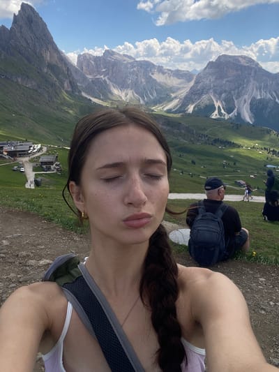 This is a selfie of a young woman with her eyes closed, taken outdoors with a mountainous landscape in the background. She has brown hair braided and is wearing a light purple tank top. The background features a steep, grassy mountain slope with scattered buildings that appear to be a small village or resort. In the mid-ground, several people are visible, some sitting on the ground, and one person is walking with a backpack. The sky is bright blue with fluffy white clouds. The overall scene suggests a daytime activity in a scenic, mountainous region. The subject's closed eyes and slightly pursed lips convey a sense of relaxation or enjoyment of the moment. The terrain is rocky and dirt path in the foreground. No text is visible in the image.