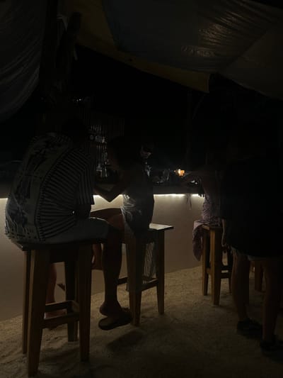 The image shows a dimly lit outdoor bar in the Cobano District of Costa Rica. Three people are seated at the bar, all of whom are partially obscured by the shadows. Two individuals are sitting on wooden bar stools facing each other, while the third person is seated to their right. They are all wearing casual attire. The bar counter is lit by a strip of LED lights. The surface behind the bar is light brown. The floor is a light tan, sandy color. The bar appears to be underneath an awning. The only light source is a string of lights above the bar, and the space is mostly dark. A low wooden partition appears behind the bar. The image's coordinates are 9.635 latitude and -85.158 longitude.