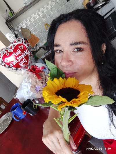 A woman poses with a sunflower, holding the stem in her left hand. On a red table, there's a plate, two mugs, a teddy bear wrapped in clear plastic with a red bow, and a "Happy Valentine's Day" heart-shaped balloon. The background shows a tiled wall and what appears to be a kitchen sink. The date "14/02/2020" and time "11:49" are visible in the lower left corner.