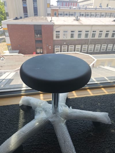 The image shows a close-up of a black, oval-shaped stool seat with a metallic base. The stool's base is partially wrapped in clear bubble wrap. The stool is placed on a dark grey carpet next to a window. Outside the window, a cityscape with multiple buildings is visible. The buildings appear to be residential or office blocks, with many windows. The lighting suggests it is daytime. The scene appears to be indoors, near a window. There is no visible text in the image. The orientation of the image is such that the stool is the primary focus in the foreground.