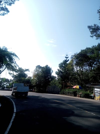 The image captures a street scene in a location with lush vegetation. A truck is parked on the left side of the road. On the right side of the road, there's a small structure with a sign written on it. A colorful umbrella stands beside it, with chairs and a small table underneath. The road is paved with a yellow line marking the center. The sky is bright and clear. There are trees and bushes surrounding the road, and in the background, a tall pine tree can be seen. The location is not explicitly identified as Phouviang-Gnai, Laos.