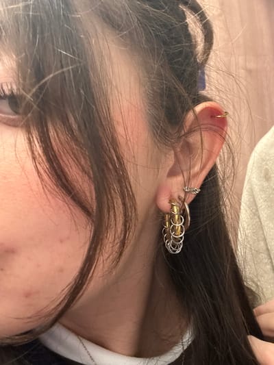 A close-up shot shows a person's ear, extensively decorated with piercings. A gold hoop is visible at the top, with a complex earring design mid-ear consisting of stacked gold and silver rings joined together, suggesting a woven or chainmail appearance. Loose, dark brown hair partially obscures the ear and face. The individual has fair skin and a glimpse of an eye is visible.