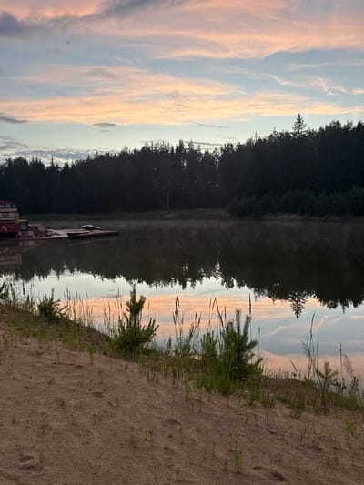 The image depicts a serene natural landscape at dusk, likely near the city of Sosnovka, Russia. A calm body of water occupies the central and lower portions of the frame, reflecting the soft hues of the sunset sky and the dark silhouette of a dense forest lining the opposite bank. Scattered on the water are a few wooden structures, including a dock with what appears to be a jet ski or personal watercraft. The foreground consists of a sandy embankment with sparse vegetation, including young pine trees and tall grasses. The sky is a blend of pale blues and soft pinks, characteristic of twilight, with wispy clouds. The overall atmosphere is tranquil and natural. No people are visible in the scene.