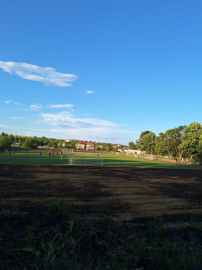 The image captures a wide-angle view of a sports complex, specifically a football pitch, situated in Ezerovo, Bulgaria, under a clear blue sky with scattered white cumulus clouds. The foreground consists of a dark, unpaved, uneven earth area showing distinct horizontal striations, with sparse green vegetation emerging from the soil.
The midground features a vibrant green grass football pitch, illuminated by late afternoon or early evening sunlight, casting long shadows. On the pitch, numerous individuals, primarily males, are actively engaged in a football game or practice. They are clad in distinct sportswear: several players in blue, some in orange, others in red, and one individual identifiable as a goalkeeper in a bright green kit positioned near the goal. A white-netted football goal stands prominently on the left side of the pitch, closer to the viewer.
Beyond the pitch, in the background, a residential area is visible, comprising various buildings with tiled roofs, some orange/red and others lighter in color. A dense line of mature green trees, particularly on the right side, separates the pitch from further structures and a hilly landscape. A dirt track or path runs alongside the pitch on the right, where a few individuals are seen walking or sitting. Utility poles and overhead lines are also present, indicating infrastructure. The overall scene suggests an active community sports facility.