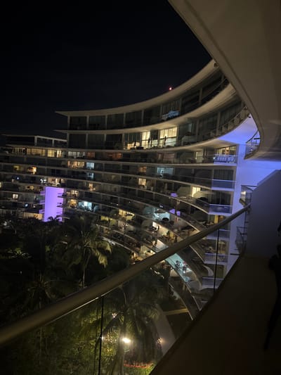 A nighttime view of a tall, curved building with multiple floors. Each level features a series of illuminated windows and balconies. Palm trees grow nearby, partially obscuring the lower floors. The architecture includes arches and support beams, visible against the dark sky. Some areas are lit with purple lighting, contrasting with the warmer light from the building's windows.
