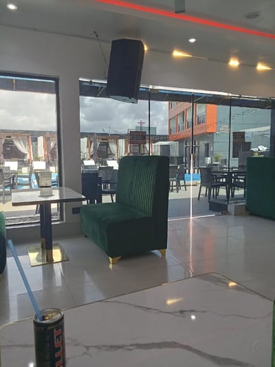 The interior of a restaurant located at the coordinates (latitude: 5.472, longitude: 5.817) in Ujevwu, Nigeria, is shown. There is a green sofa with gold legs in the foreground, along with a small table with gold legs, and a beverage can on the table. The window behind the table looks out into the courtyard of a hotel, which features a swimming pool and patio furniture. The restaurant is brightly lit, with a speaker hanging from the ceiling and a red light strip running along the ceiling. The walls are white, and the floor is tiled. There are two windows in the room, looking out on a courtyard with a pool. There is outdoor furniture in the courtyard. There is a second, dark green sofa in the background. There are no people present. The scene suggests a restaurant that is likely open to the public and is intended for casual dining. The image appears to have been taken during the day, as the sun is shining outside. There is a slight glare on the floor in the center of the image.