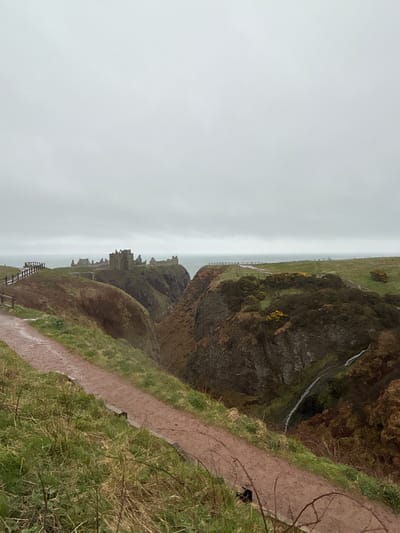 The image depicts the Dunnottar Castle in Stonehaven, United Kingdom. The castle ruins are situated on a rocky headland jutting into the North Sea. A narrow, muddy footpath runs along the edge of a cliff towards the castle. The sky is overcast and grey. A waterfall flows from the cliff edge. A wooden fence is visible along the path. The vegetation consists of grass and bushes, showing signs of dampness.