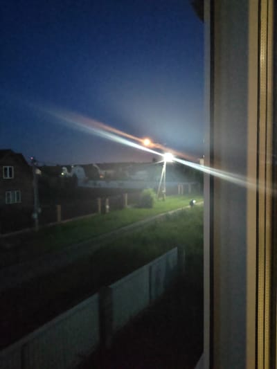 The image shows a night view from a window, looking out onto a residential area in Zhal'skoe, Russia. The sky is dark blue, transitioning to black towards the top. A bright streetlight illuminates the scene, creating a strong lens flare that streaks across the sky and a portion of the landscape. In the mid-ground, there are several houses with visible windows and roofs. A dark-colored car is parked on the side of a pathway. The foreground features a concrete fence and a grassy area. The right side of the image is dominated by the window frame, with vertical blinds visible. The overall impression is a quiet, possibly rural or suburban setting at night.