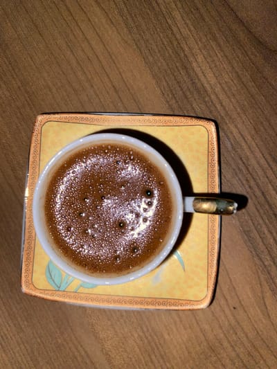 A close-up, top-down shot shows a white cup filled with dark, frothy coffee. The coffee has a foamy top with small bubbles and shimmering highlights, indicating it's freshly brewed. The cup has a small, ornate gold handle. It sits on a decorative square saucer with a yellow background, featuring a repeating scroll pattern around the edge and a subtle floral design in the center. The saucer and cup are placed on a wooden surface with a visible grain, suggesting a table or desk. The lighting casts a slight shadow from the cup and handle onto the saucer, implying the light source is from above and slightly to the right. The scene is static, with no people or discernible action, and the overall mood is peaceful and inviting, suggesting a moment of quiet enjoyment. Given the context of Kocasinan, Turkey, this could be a depiction of Turkish coffee, a culturally significant beverage often enjoyed in a relaxed setting. The time of day and weather are not discernible from the image. There is no visible text in the image.