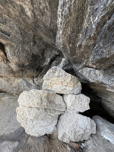 This is a close-up, eye-level, outdoor, medium shot of a pile of weathered, light gray boulders stacked in the foreground, nestled against a dark gray, rocky overhang that recedes into shadow. The boulders are rough and granular in texture, with visible cracks and indentations. The rock formation is substantial, with large, angular segments creating a sense of depth and cavernous space. The environment suggests a natural, possibly coastal or mountainous, setting, with some dry dirt and debris visible at the base of the rocks. The lighting is natural, creating distinct shadows and highlighting the varied textures of the stone. There are no people or animals visible in the image. This scene is located at Dom Otdykha Zolotoy Plyazh, Russia.