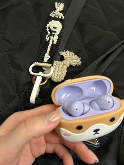 A person is holding an open, decorative earbud case. The case is a light tan color with a white section showing the face of an animal; there are black dots and a black symbol where the animal's face would be. Inside the case are two lavender earbuds. The person's hand is fair-skinned with long, clear nails. The background appears to be a black, quilted jacket with a black strap. There is a crocheted keychain attached to the strap, showing a guitar. The location is unknown.