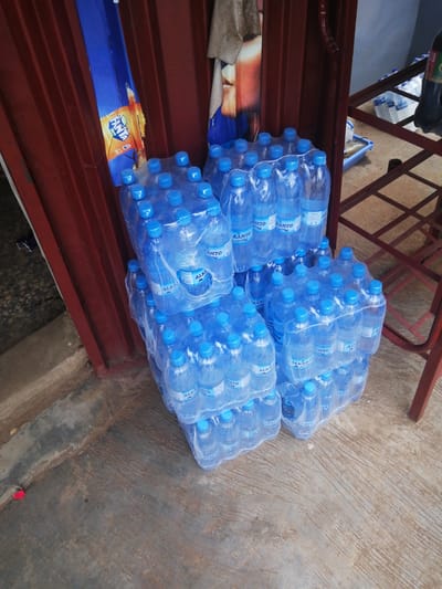 The image shows a stack of bottled water arranged in packs of six, wrapped in clear plastic. The bottles have blue caps and labels that read "ALKAMTO". The packs are placed on a concrete surface, next to a red metal structure that appears to be part of a stall or display. Behind the water bottles, a red metal door with vertical lines is visible, partially obscured by a blue and orange banner advertising "FANTA". The banner has an image of an orange and the Fanta logo. The overall setting suggests an outdoor market or shop in Ajasse Ipo, Nigeria. The lighting indicates it is daytime, and the ground appears somewhat weathered, suggesting an environment exposed to the elements. There are no people visible in the image. The focus is on the stacked merchandise, likely for sale.