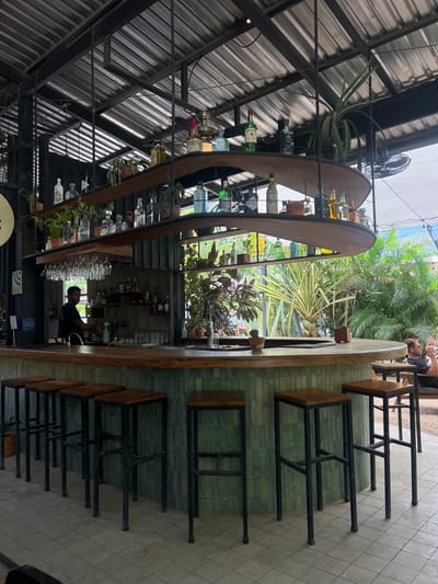 The image shows an outdoor bar area in Santa Fe, Costa Rica, located at coordinates 9.642 latitude and -85.165 longitude. The bar is a modern, curved design with a green tile facade and a wooden countertop. The bar is equipped with a metal shelving system behind it displaying various bottles and glasses. Several black metal bar stools are arranged around the bar. A man is standing behind the bar, facing left, presumably preparing drinks. The bar is situated in a partially enclosed space, with lush tropical foliage visible behind it. The environment is open-air, with a metal roof structure overhead. The bar is lit with overhead lighting and the scene is bright and well lit. The image suggests a lively, tropical atmosphere with a modern design aesthetic.