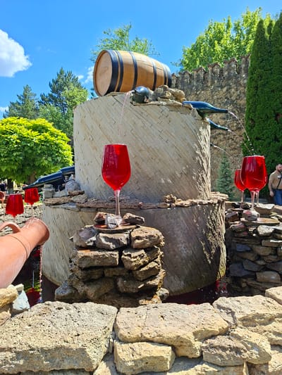 The image depicts an outdoor wine-themed fountain located in Milestii Mici, Moldova. The fountain is designed to resemble a wine barrel and its surroundings, with elements such as large wine glasses filled with red liquid, wine bottles, and a wooden barrel. Water or wine flows from the bottles into the glasses and also cascades down the stone structure. The fountain is surrounded by natural landscaping, including trees and a stone wall. Several large wine glasses are prominently featured, with some appearing to be overflowing. A clay jug is also visible, positioned to pour liquid into the fountain's basin. The scene is set under a bright, sunny sky with scattered clouds. A man in a light-colored shirt and dark pants is visible on the right side of the image, appearing to observe the fountain. The overall impression is that of a decorative and whimsical display related to wine production and consumption.