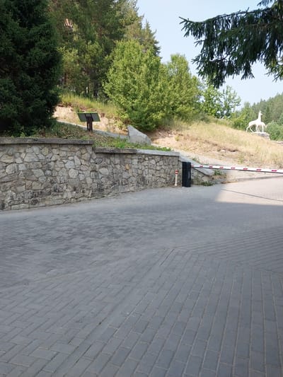 This image depicts an outdoor scene with a paved walkway in the foreground. To the left, there is a stone wall with greenery growing above it. Beyond the wall, a wooded hillside is visible with several trees, and on the top of the hill, a white statue of a deer is present. In the middle ground, a barrier arm is extended across the walkway, suggesting an entrance or checkpoint. The environment appears to be a park or recreational area. The image is likely taken in the city of Belokurikha, Russia, known for its natural beauty and resorts.