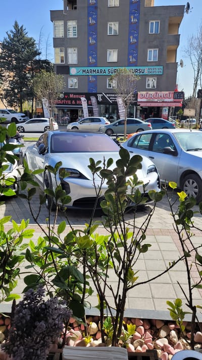 The image captures a street scene in Beylikdüzü, Turkey. A line of parked vehicles occupies the foreground, partially obscured by foliage. The vehicles are various shades of white and silver. A grey building is visible in the background, featuring a blue, nautical-themed mural on its side. Several signs are attached to the building, including "MARMARA SÜR KURSU" (a driving school) and "DÜRÜMCÜ" (a type of Turkish wrap). Other businesses like a hairdresser are present. The sky is light blue and clear. The image appears to be taken at ground level.
