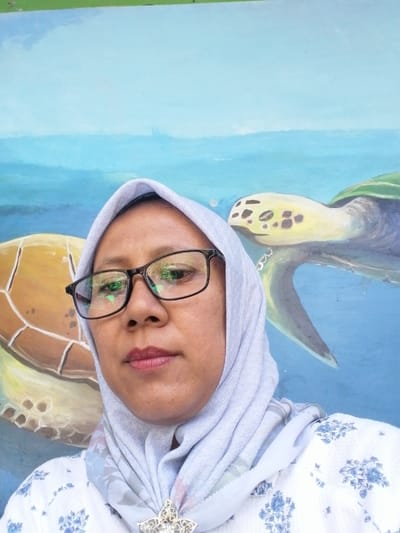 In Tegalwangi, Indonesia, a woman wearing a light blue hijab and a white patterned top with blue flowers is taking a selfie in front of a mural. The mural depicts two sea turtles swimming in blue water. The woman is wearing dark-rimmed glasses, and her face is the primary subject of the photograph. A decorative brooch with rhinestones is attached to her hijab. The lighting suggests it is daytime, and the environment appears to be outdoors or in a well-lit open space.