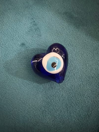 A heart-shaped, deep blue glass object, designed to resemble an evil eye, rests on a textured, teal-colored surface. The object has a prominent eye motif in the center, composed of concentric circles of white, light blue, and black. The lighting creates a slight shadow and highlights on the glossy surface of the glass. The scene is devoid of human presence and does not provide any contextual information that would allow for its location to be specified within the city of İzmir, Turkey, or any other urban environment.