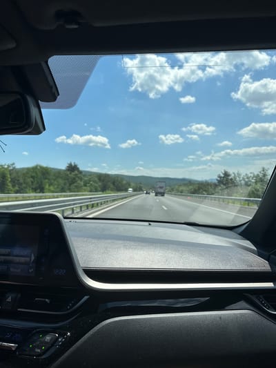 The image is taken from inside a vehicle, looking out through the windshield at a highway. The dashboard of the car is visible in the foreground, including a screen displaying the time as 12:22. The road ahead is lined with a metal guardrail and bordered by lush green trees and hills. The sky is bright blue with scattered white clouds, suggesting a sunny day. There are other vehicles on the highway, including a truck in the distance. The image does not provide specific information about the city of Rajana, Bulgaria, but depicts a typical highway scene consistent with travel in a rural or semi-rural area.