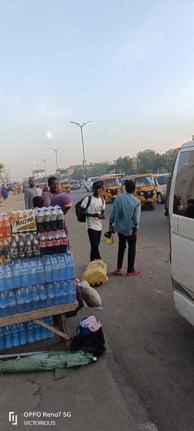 This photograph captures a vibrant street scene in Gyadi Gyadi Arewa, Nigeria, under a clear, bright sky, suggesting daytime, likely morning or late afternoon.
In the foreground to the left, a wooden vendor's stall is laden with various bottled beverages. Brands such as "MALTINA," "PEPSI," "COCA COLA," and numerous packs of "ARA" bottled water are prominently displayed. Behind the stall, several individuals are visible: a man wearing a traditional cap, another smiling man looking towards the camera, and a person with a headscarf partially obscured by the drinks.
In the mid-ground, two men stand on the paved sidewalk. One, wearing a white and green t-shirt with a backpack, is engrossed in his phone. The other, in a light blue shirt and dark trousers, sporting red flip-flops, holds a small yellow bag and faces away from the camera. At their feet are two large bags, one yellow and one off-white, along with a rolled-up green tarp or umbrella.
The background features a busy road with multiple yellow three-wheeled auto-rickshaws, commonly known as Keke Napep, indicating active traffic. Tall streetlights with distinctive Y-shaped tops line the street, leading into the distance where buildings and trees are visible. A white minivan is partially seen on the far right.
The image depicts a slice of daily life in a bustling Nigerian urban setting, showcasing street vending, pedestrian movement, and local transportation. A visible watermark at the bottom left reads "OPPO Reno7 5G VICTORIOUS."