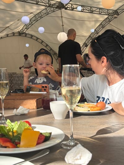 In Morozovo, Russia, at an outdoor dining event under a large tent structure with decorative spheres hanging from the ceiling, a young boy wearing a black baseball cap is being fed by a woman with dark hair. The boy is sitting at a table with a smartphone and a wooden box in front of him. A glass of champagne sits in the foreground, as does a plate of salad. In the background, other people are visible, including a man in a black t-shirt. The overall scene depicts a casual meal in a pleasant outdoor setting.