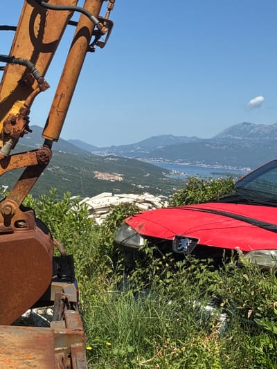 In the city of Prijeradi, Montenegro, an excavator arm and bucket, rusted and weathered, dominate the left side of the frame. The arm extends diagonally upwards, casting a shadow on itself. In the foreground, overgrown green vegetation envelops a partially visible red car, its hood and front bumper peeking through. The car's emblem, a stylized lion, is visible. Beyond the vegetation and the car, a scenic vista unfolds. Rolling green hills dotted with buildings descend towards a sparkling blue bay, a town or city nestled along its shores. Distant, hazy mountains form the backdrop, with a few white clouds scattered across the clear blue sky. The scene appears to be an outdoor, natural environment, possibly a junkyard or an abandoned site overlooking a coastal town. No people are visible in the image.