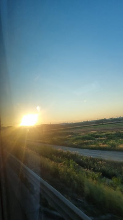 The image captures a sunset viewed from what appears to be a moving vehicle, likely a train or car, with a reflection visible on the window. The sun is a bright, dominant light source on the left side of the frame, casting long rays across a rural landscape. The foreground is occupied by a dark, reflective surface, possibly the interior of the vehicle and a portion of its structure, like a window frame or guardrail. Beyond this, a dirt or gravel road is visible, bordered by fields of green grass and some scattered vegetation. In the distance, the landscape appears to be agricultural land with some man-made structures, possibly buildings or storage facilities, along a gentle rise. The sky transitions from a deep blue at the top to a pale yellow and orange hue near the horizon, indicating the time of day is late afternoon or early evening. While no specific landmarks are identifiable, the scene suggests a semi-rural or agricultural setting. The location is specified as Nicolae Bălcescu, Romania, which is a commune in the Constanța County.