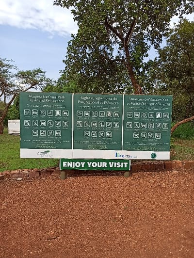 The image depicts a sign at Akagera National Park displaying park rules and regulations in English, French, and Kinyarwanda. Various symbols are used to illustrate the regulations. A separate sign encourages visitors to "ENJOY YOUR VISIT" in green letters. The area surrounding the signs appears to be a pathway covered with reddish-brown gravel. Trees and vegetation are visible in the background.