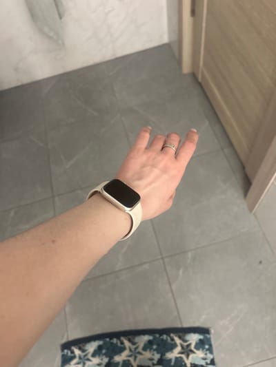 The image shows a close-up view of a person's left arm and hand, wearing a white smartwatch and a silver ring. The arm is positioned horizontally across the frame, with the smartwatch on the wrist and the hand extended forward. The background consists of a tiled floor with a subtle marble pattern, and a wooden doorframe to the right. The scene appears to be indoors, possibly in a bathroom or hallway. There is no information available to determine the location in the city of Нижние Муллы, Russia.
