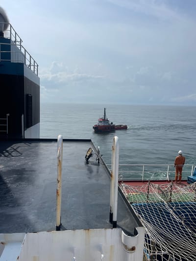 The scene, located in the city of Orimedu, Nigeria, is captured from the deck of a large vessel overlooking open water. In the foreground, a dark grey deck, appearing damp, features two prominent, partially rusted white stanchions and an upward-angled floodlight. A black and blue superstructure with white railings occupies the far left. On the vessel's starboard side, a person in an orange jumpsuit and white hard hat stands facing away from the camera, observing the sea. A red and blue safety net is visible below this individual. In the mid-distance, a red and white tugboat, characterized by its superstructure and mast, moves leftward across the calm, grey-green water, generating a visible wake. The sky above is largely overcast with scattered clouds.