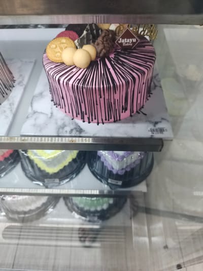 The image shows a display case filled with cakes. In the foreground, there are several small, heart-shaped cakes in plastic containers, with varying frosting colors including white, yellow, and purple. Above these, a larger, round pink cake with black drizzles is prominently displayed. This cake is decorated with cookies, small round sweets, and a "Jatayu Bakery" logo. A partially visible "Happy Birthday" topper is also present. Another similar pink cake is partially visible to the left. The display case is made of metal and glass. The background shows the interior of what appears to be a bakery. The image was taken in Krian, Indonesia, at Jatayu Bakery.