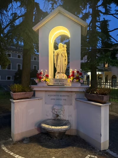 The image depicts a religious shrine at dusk. The shrine, constructed of white stone, features a statue of the Virgin Mary and an inscription "AVE MARIA GRATIA PLENA." Floral arrangements adorn the shrine at two levels. Adjacent to the shrine is a stone drinking fountain with a decorative lion's head. The plaque on the shrine mentions "ACQUEDOTTO MARIOTTO ANNO SANTO 1950." The environment includes trees, a building in the background, and cobblestone pavement. The overall location is identifiable as Villafranca di Verona, Italy.