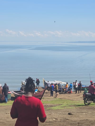 The scene is a lakeside landing in Homa Bay, Kenya. The latitude and longitude provided ( -0.522, 34.459) correspond to the location of Homa Bay. The water of Lake Victoria is in the background. The sky is a clear blue with a few white wispy clouds. The foreground consists of a dirt shoreline, a group of people, and several boats. A few people are standing on a wooden dock, while others are walking toward or away from the boats. A man is seated on a small motorcycle. The boats are tied to the dock and are either empty or have a few people inside. There is a small, red boat with a sign reading "HOMUWENDO".